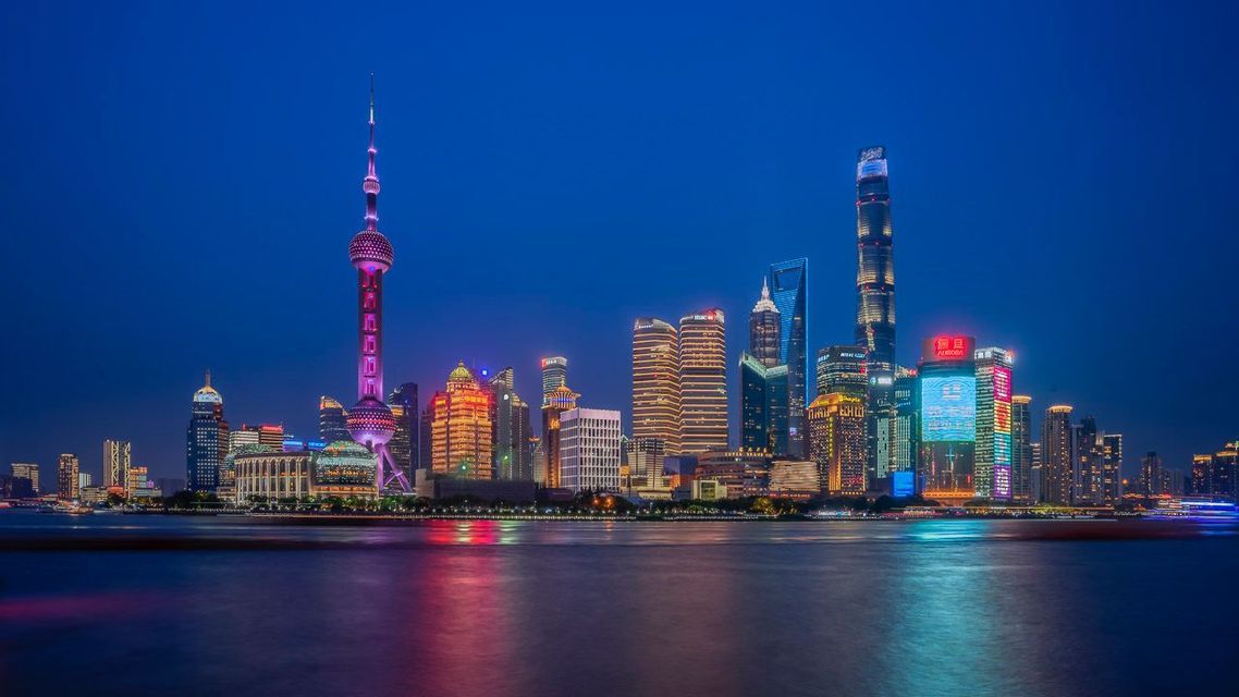 A modern city skyline at night, with illuminated buildings and a tall tower reflecting on the calm water.