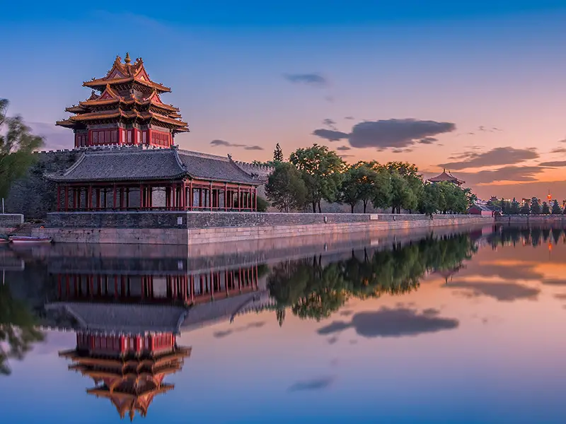 Une tour d'angle chinoise traditionnelle sur un mur en pierre se reflète dans l'eau calme des douves au coucher du soleil.