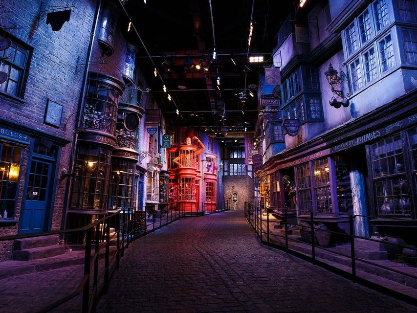Una strada acciottolata serpeggiante su un set cinematografico, costeggiata da negozi fantastici e storti sotto luci viola e blu suggestive.