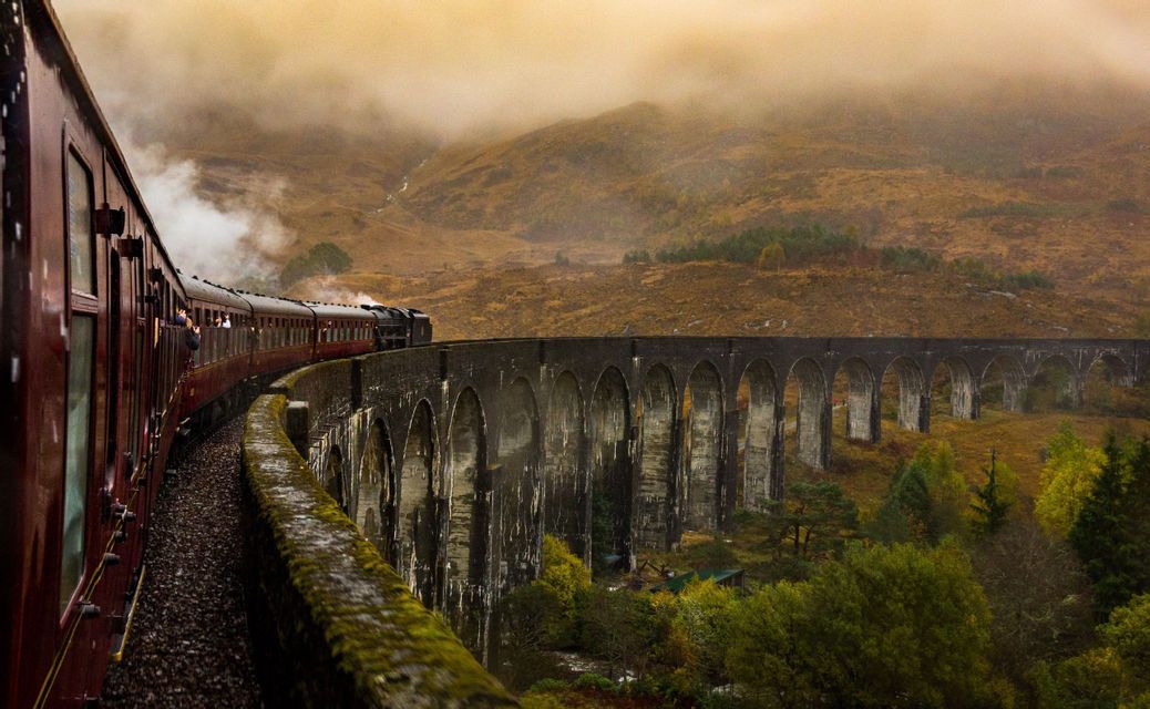 Un treno a vapore rosso attraversa un lungo viadotto in pietra ad arco in un paesaggio nebbioso e autunnale.