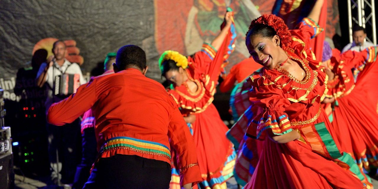 Ballerine in colorati costumi tradizionali rossi si esibiscono su un palco, con una donna sorridente in primo piano.