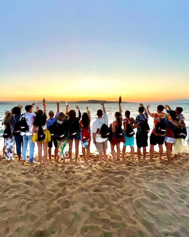 Un viaje en grupo de WeRoad, visto por detrás, de pie en una playa de arena con los brazos en alto, observando un atardecer sobre el mar.