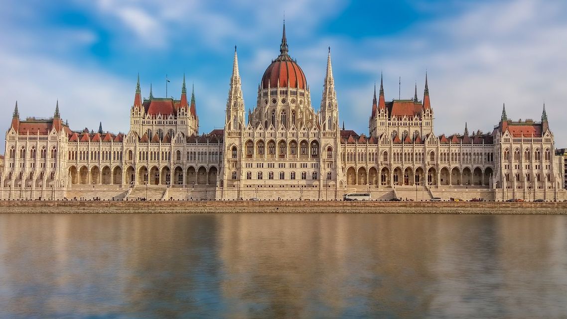 Un gran y ornamentado edificio gubernamental con una cúpula roja central y agujas se refleja en un ancho río bajo un cielo azul.