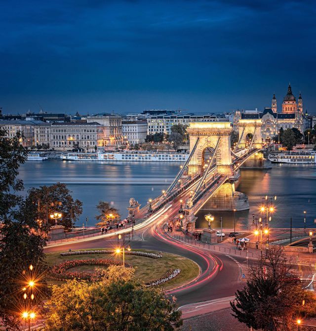Un ponte illuminato su un fiume al tramonto, con le scie luminose del traffico che portano a una città con edifici storici.