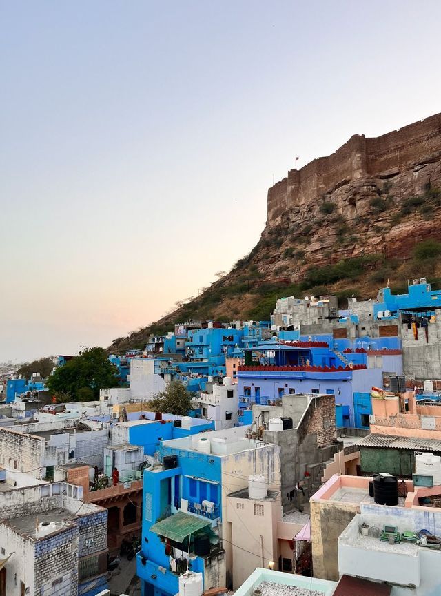 Un fitto paesaggio urbano di edifici bianchi e blu, raggruppati ai piedi di una grande collina rocciosa sormontata da una fortezza in pietra al tramonto.