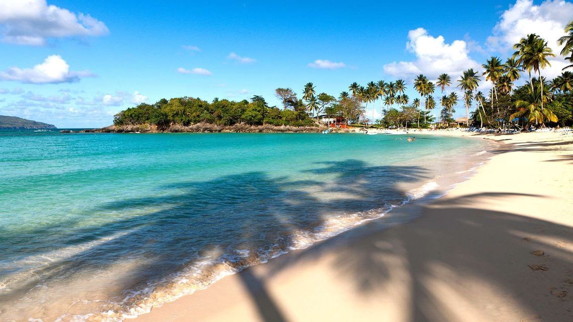 Ombre di palme cadono su una spiaggia di sabbia bianca accanto ad acqua turchese, con una lussureggiante penisola verde sullo sfondo sotto un cielo blu.