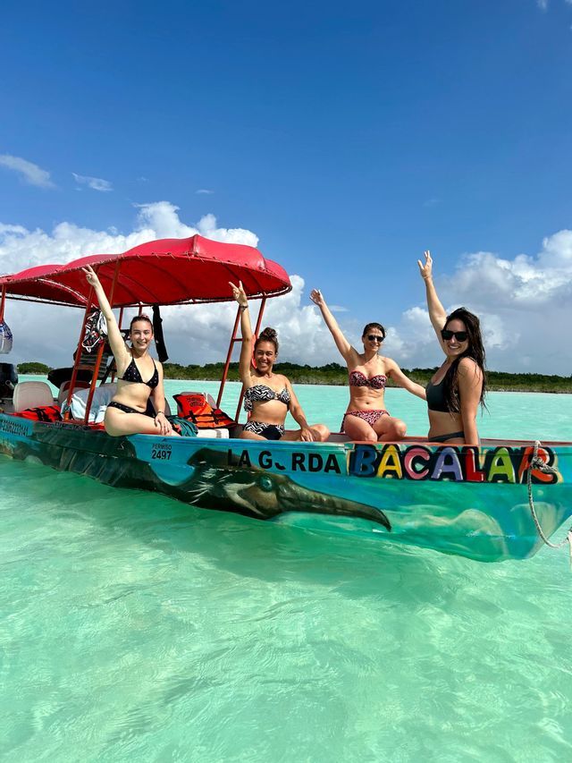 Vier Frauen in Bikinis auf einer WeRoad-Gruppenreise lächeln mit erhobenen Armen auf einem Boot in türkisfarbenem Wasser unter blauem Himmel.