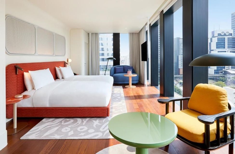 A sunlit, modern hotel room with two beds, colorful chairs, and floor-to-ceiling windows showing a cityscape.