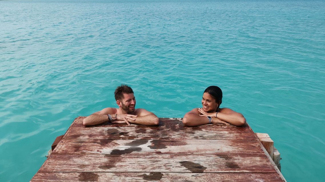 A man and a woman on a WeRoad group trip lean on a wooden dock, smiling at each other in turquoise water.