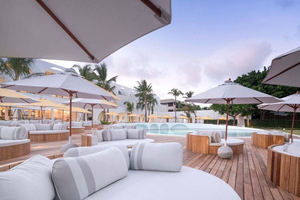 An outdoor lounge area at a resort with white daybeds and umbrellas on a wooden deck by a swimming pool.