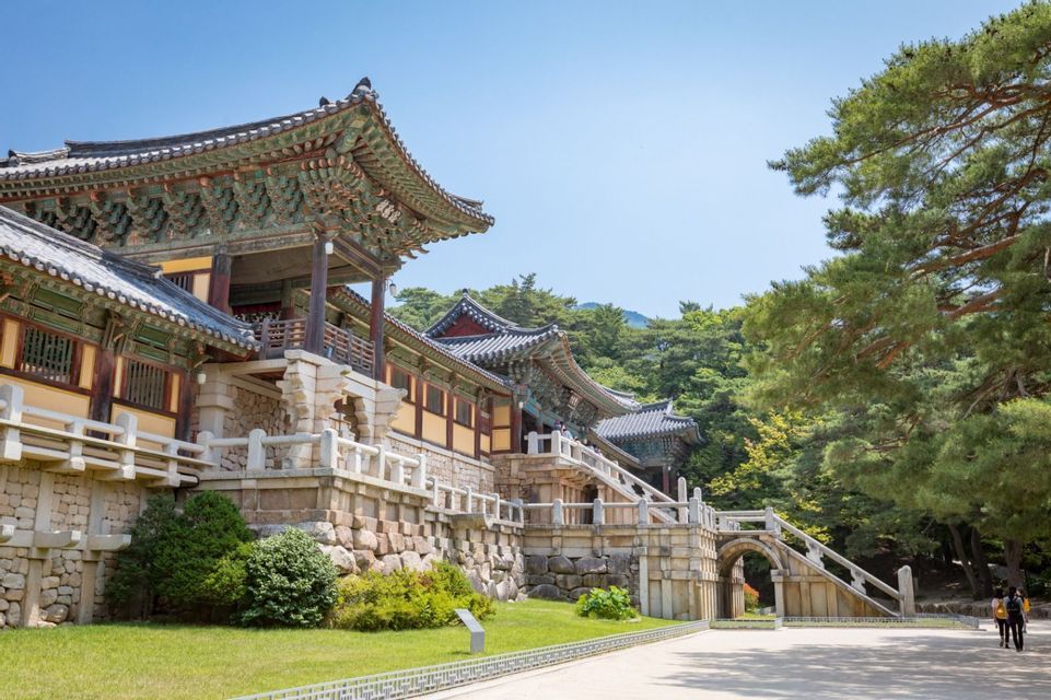 Ein traditioneller koreanischer Tempel mit verzierten, gestuften Dächern steht auf einem Steinfundament, umgeben von üppigen Bäumen unter einem klaren blauen Himmel.