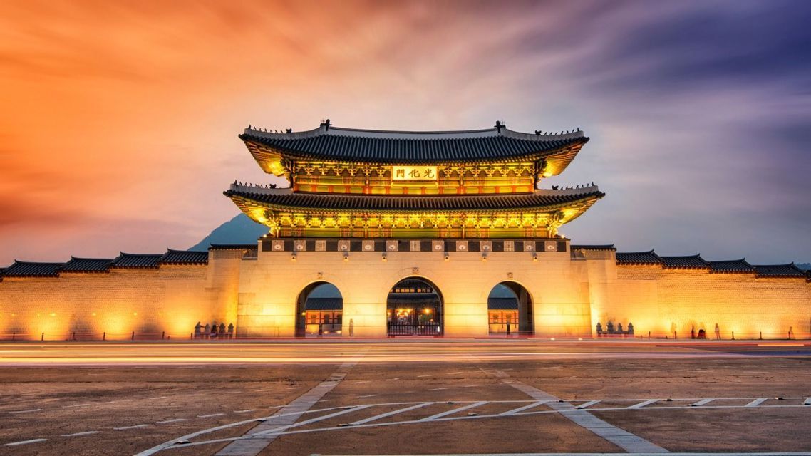 A traditional Asian gatehouse and its stone wall are brightly illuminated at dusk, with long streaks of traffic lights in the foreground.