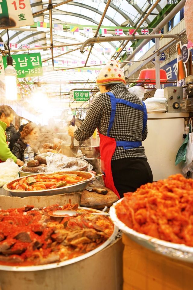 A vendor wearing a colorful beanie and apron cooks at a busy food stall in an indoor market, surrounded by large steaming bowls of food.