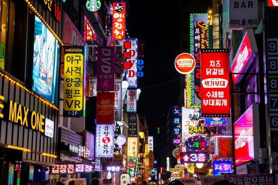 A narrow city street at night is brightly illuminated by a dense wall of colorful neon signs, mostly in Korean script.