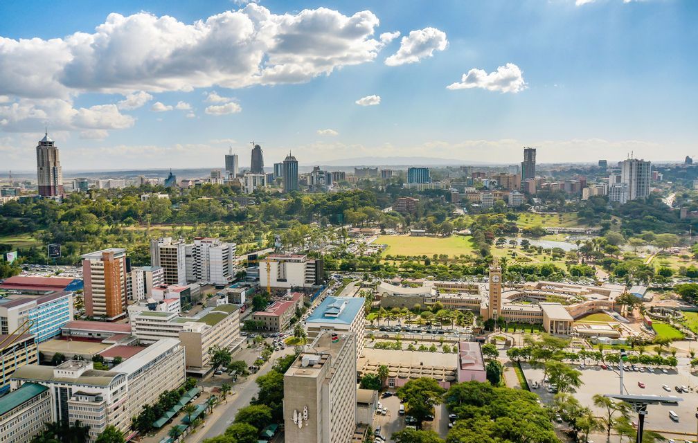 Luftaufnahme einer weitläufigen Stadtlandschaft mit modernen Wolkenkratzern und üppigen grünen Parks unter einem teilweise bewölkten blauen Himmel.