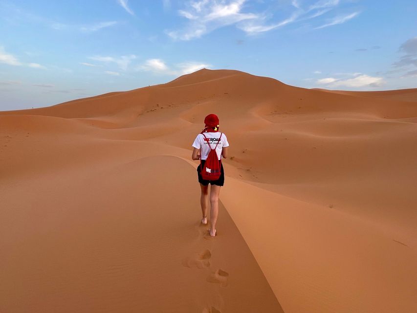 A person seen from behind wearing a WeRoad t-shirt and red cap walks barefoot up a large orange sand dune.