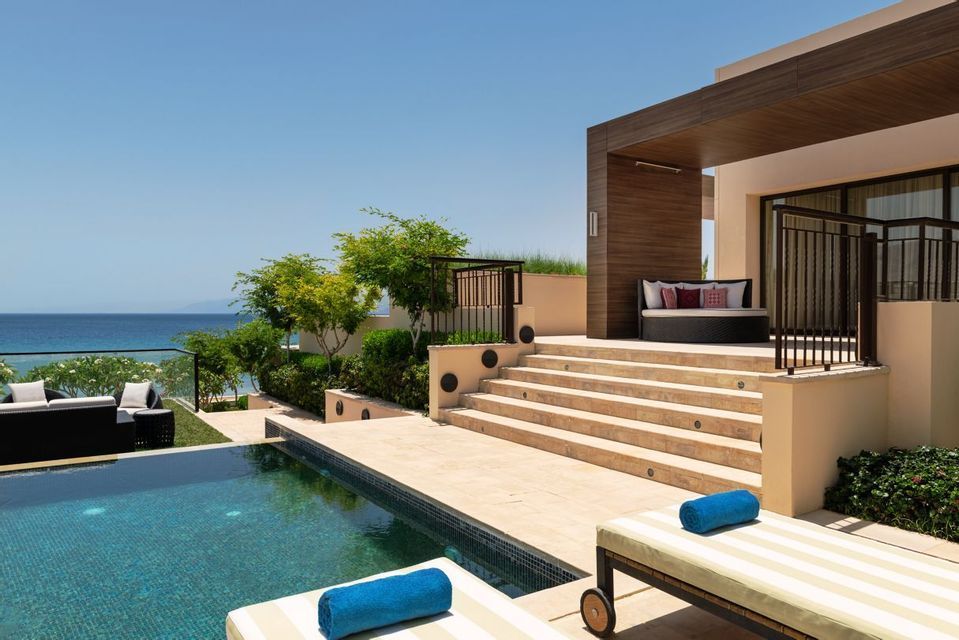 Ein moderner Hotel-Patio mit Swimmingpool und Sonnenliegen überblickt das ruhige blaue Meer unter einem klaren Himmel.
