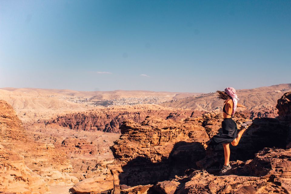 Une femme voilée pose sur une jambe sur un piton rocheux, dominant un vaste canyon désertique sous un ciel bleu clair.