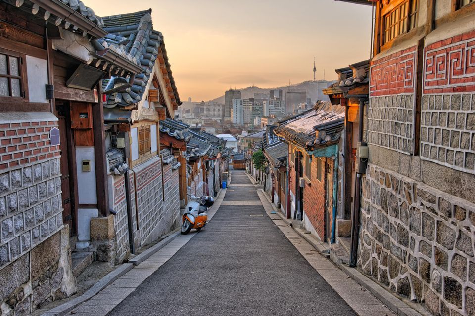 Una calle estrecha desciende entre casas tradicionales con tejados de tejas, revelando un horizonte moderno de la ciudad en la distancia al atardecer.