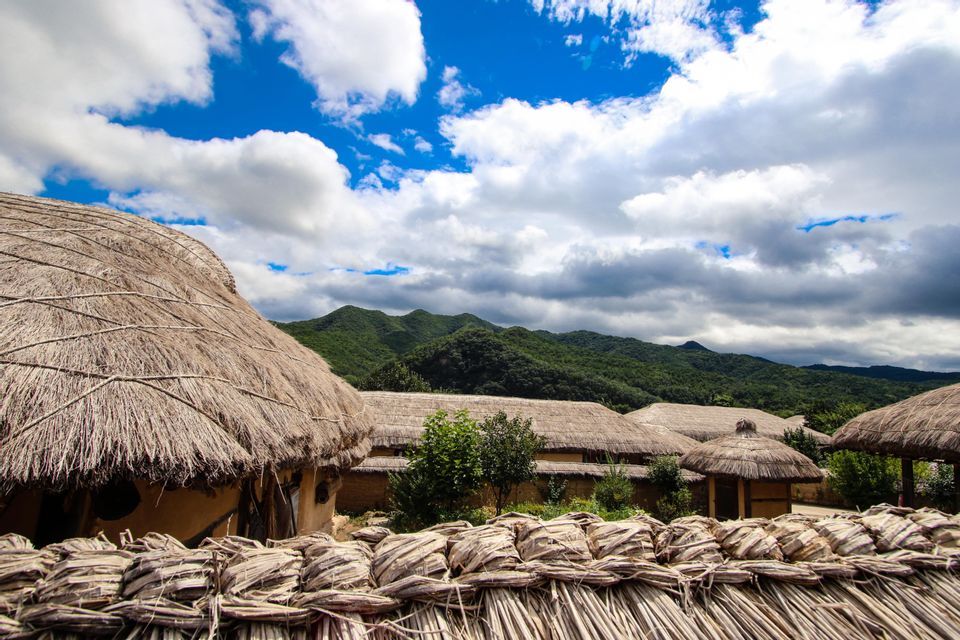 A village of traditional huts with thatched roofs sits at the base of green mountains under a partly cloudy sky.