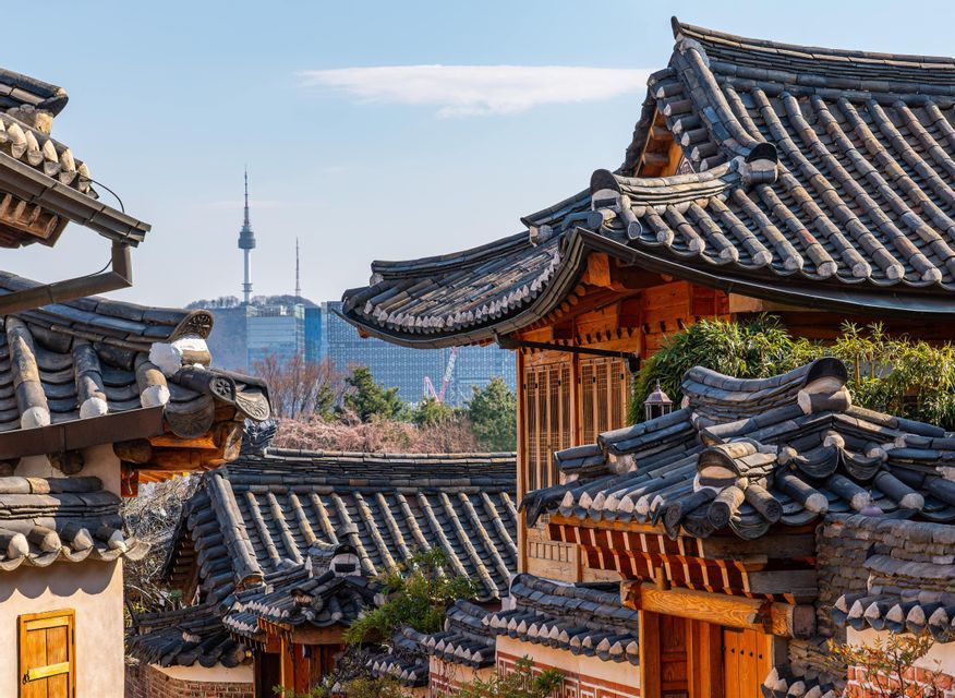 Ein Blick über traditionelle koreanische Ziegeldächer mit einer modernen Stadtkulisse und einem in der Ferne sichtbaren Kommunikationsturm.