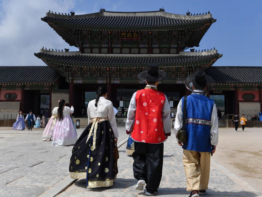 Vue de dos, trois personnes en tenues coréennes traditionnelles et colorées se dirigent vers la porte d'un palais historique sous un ciel dégagé.