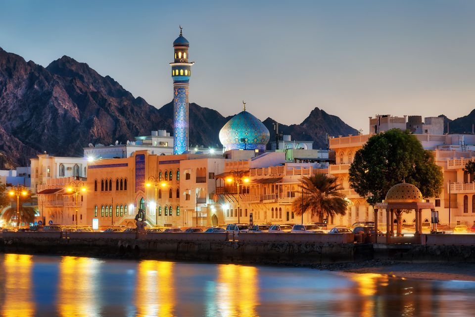 Une mosquée illuminée avec un dôme en mosaïque bleue et un minaret sur un front de mer, avec des montagnes derrière la ville au crépuscule.