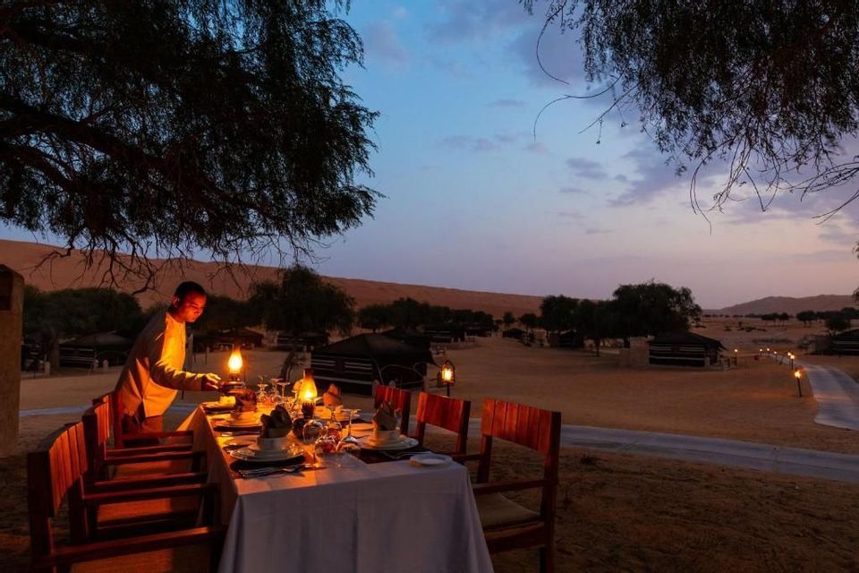 Un cameriere accende una lanterna su un tavolo apparecchiato per una cena all'aperto in un campo nel deserto durante il crepuscolo.