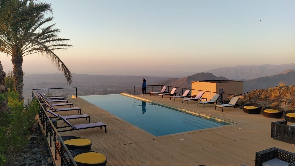 A woman stands by an infinity pool on a wooden deck, looking out over a mountainous valley at sunset.