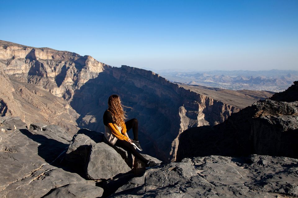A woman with long hair sits on a rocky ledge overlooking a vast, sunlit canyon under a clear blue sky.