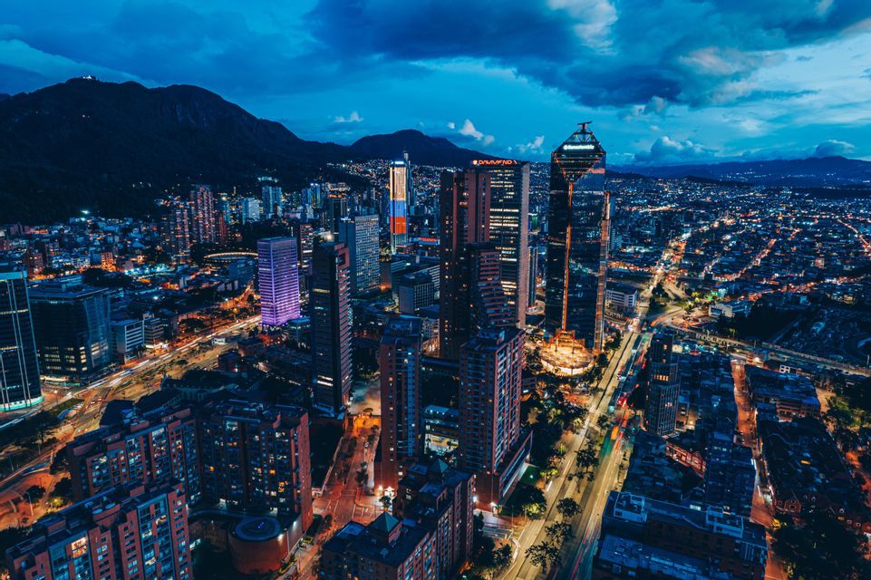 An aerial view of a sprawling cityscape illuminated by lights at dusk, with dark mountains silhouetted against a cloudy sky.