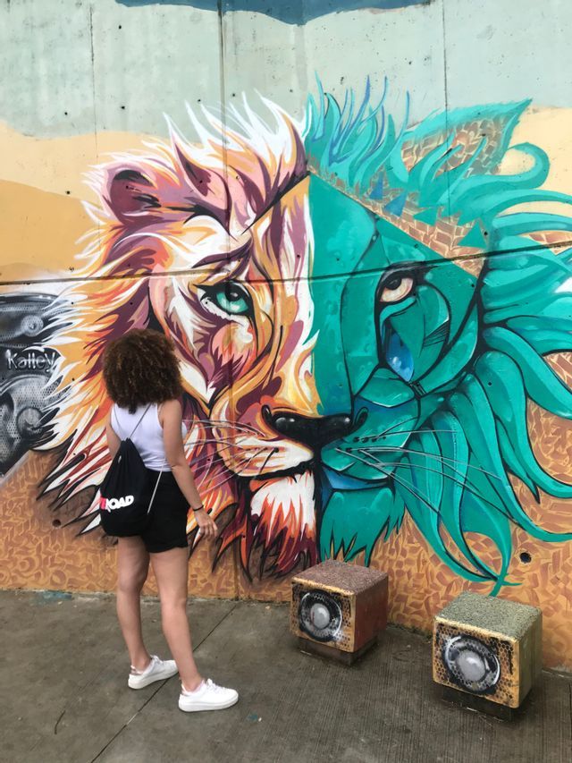 Une femme avec un sac à dos WeRoad contemple une fresque de street art colorée, un visage de lion, sur un mur en béton.
