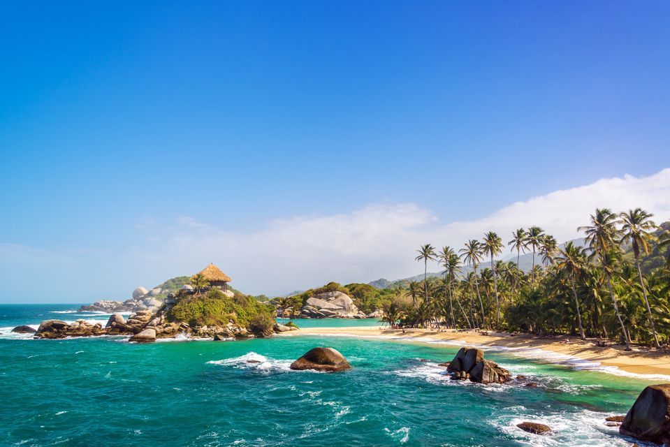 A hut with a thatched roof sits on a rocky outcrop in turquoise waters, next to a sandy beach lined with palm trees under a blue sky.