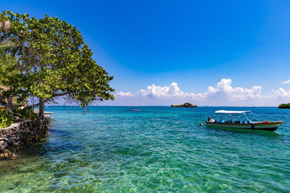 Ein Boot mit Sonnensegel liegt in klarem, türkisfarbenem Wasser an einer felsigen Küste mit einem üppig grünen Baum unter blauem Himmel.