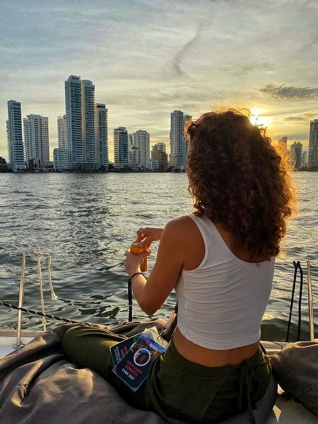 Una donna dai capelli ricci, in viaggio di gruppo WeRoad, è seduta su una barca, mentre osserva il tramonto dietro lo skyline di una città moderna oltre l'acqua.