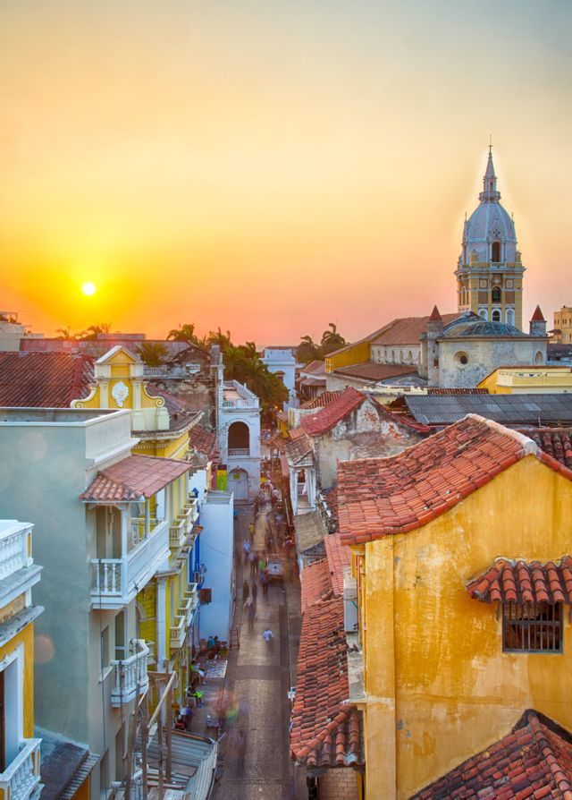 A high-angle view of a city street with colorful buildings and red-tiled roofs at sunset, with a cathedral in the background.
