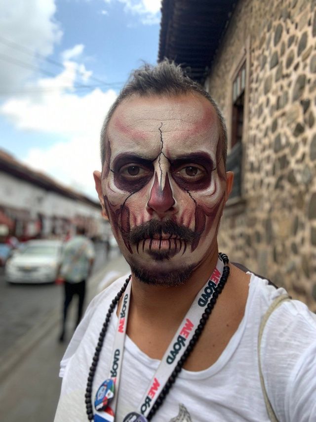 Un hombre con maquillaje detallado de calavera y un cordón de WeRoad posa para una selfie en una calle adoquinada.