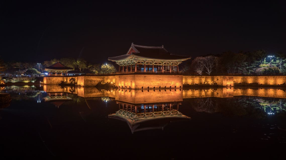 Ein traditioneller Pavillon und eine Steinmauer, nachts beleuchtet, werfen eine perfekte Spiegelung auf das ruhige Wasser darunter.