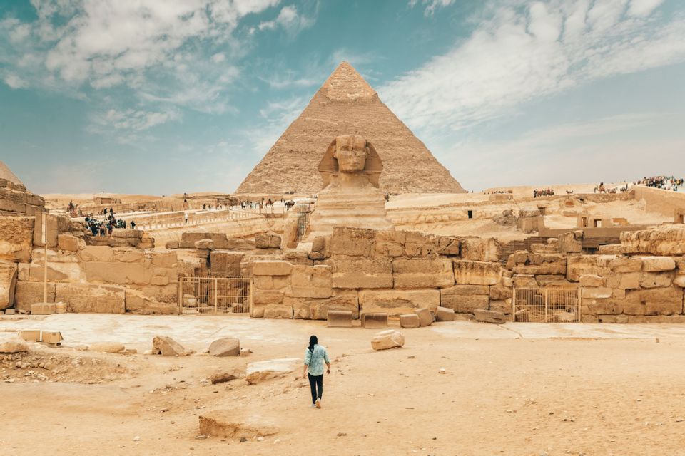 Une personne marche vers le Grand Sphinx et une grande pyramide à travers une étendue sablonneuse de ruines antiques sous un ciel partiellement nuageux.
