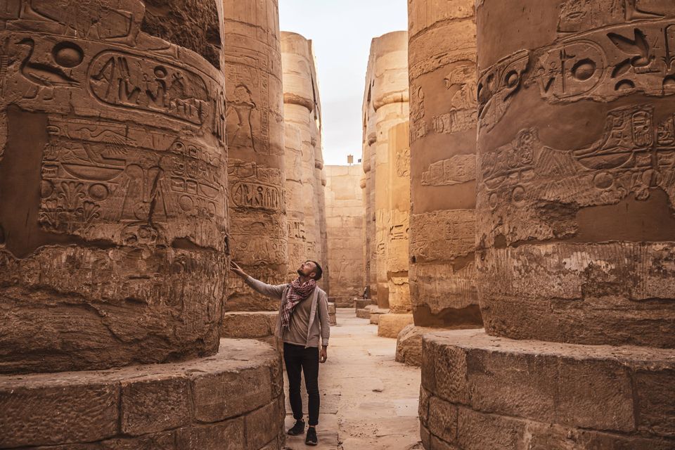 Un hombre está de pie en la sala de un templo antiguo, observando los jeroglíficos tallados en las enormes columnas de piedra.