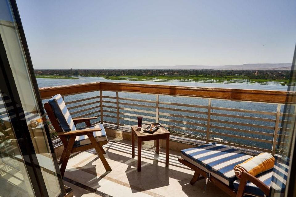 Un balcon ensoleillé avec des meubles de salon rayés et une petite table, offrant une vue sur une large rivière et des berges verdoyantes lointaines.
