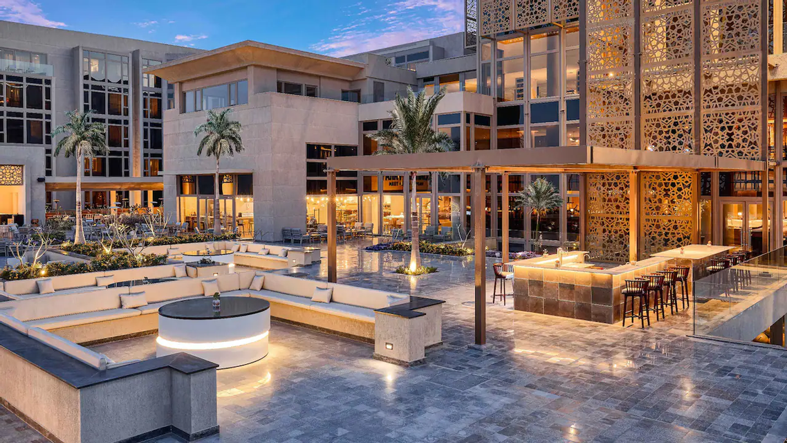 Une terrasse extérieure d'un hôtel moderne au crépuscule, avec des zones de sièges éclairées, un bar et des palmiers.