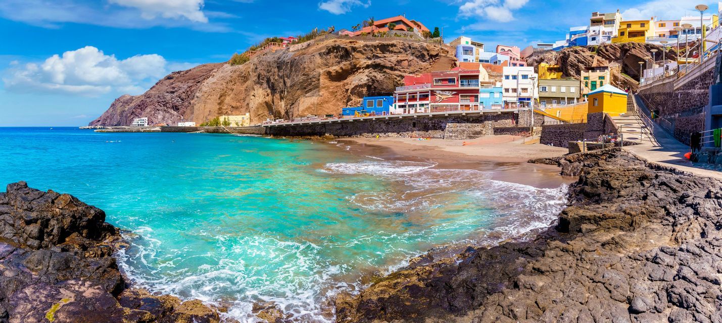 A panoramic view of a coastal village with colorful houses built into a cliff above a turquoise sea and sandy beach.