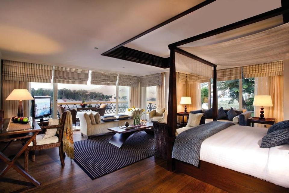 Eine Hotelsuite mit Himmelbett, Wohnbereich und großen Fenstern mit Blick auf einen Balkon und einen Fluss.