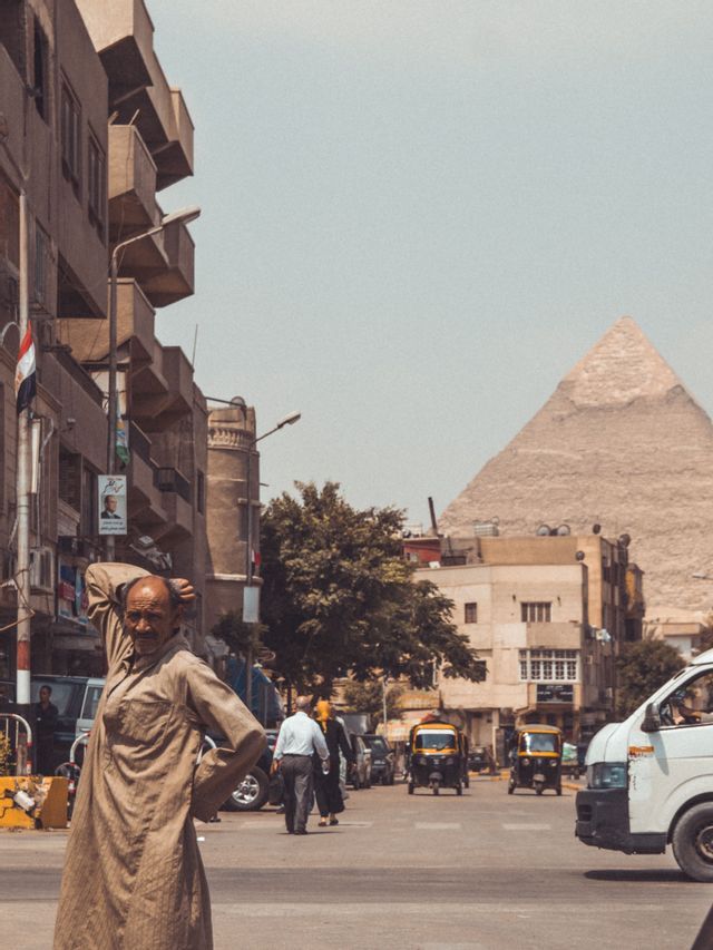 Ein Mann in einer hellbraunen Tunika steht auf einer belebten Straße, mit Verkehr, Gebäuden und einer großen Pyramide im Hintergrund.