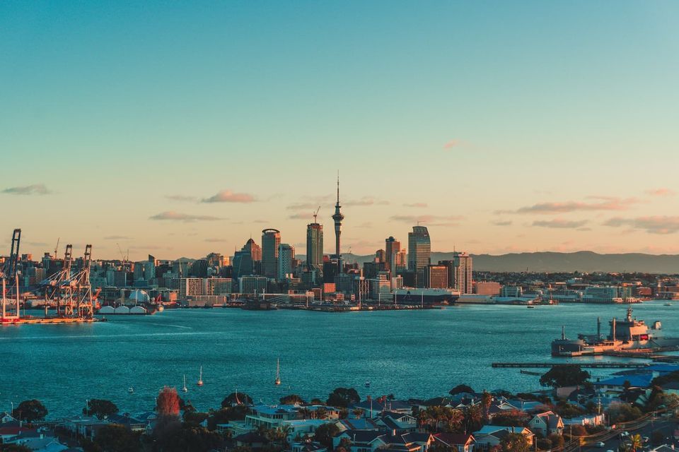 A city skyline with a tall tower sits on the edge of a harbor during sunset, with calm blue water in the foreground.