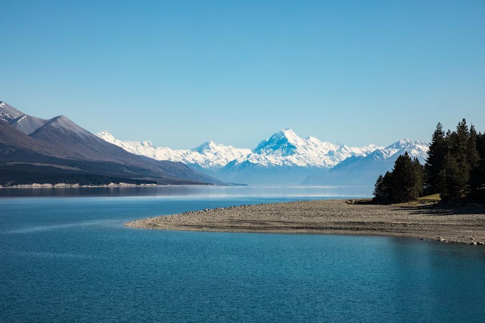 Un lac bleu et calme avec une plage de galets au premier plan, et une chaîne de montagnes enneigées en arrière-plan sous un ciel dégagé.