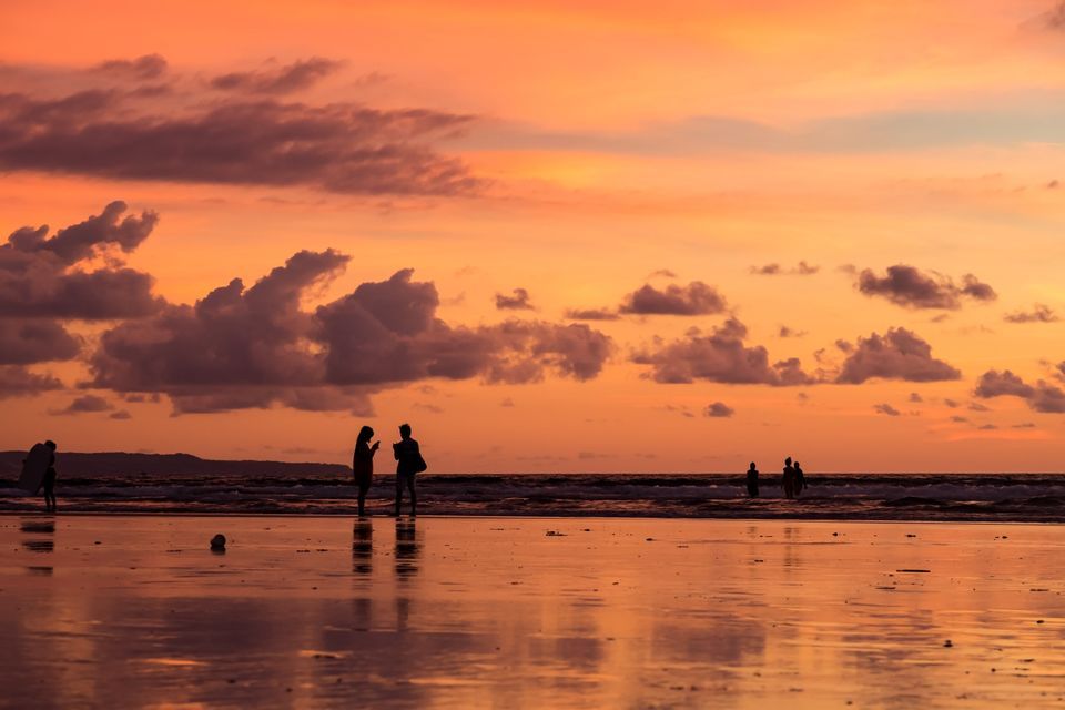 Silhouettes d'un voyage en groupe WeRoad sur une plage mouillée au coucher du soleil, avec un ciel orange et des nuages se reflétant dans le sable.