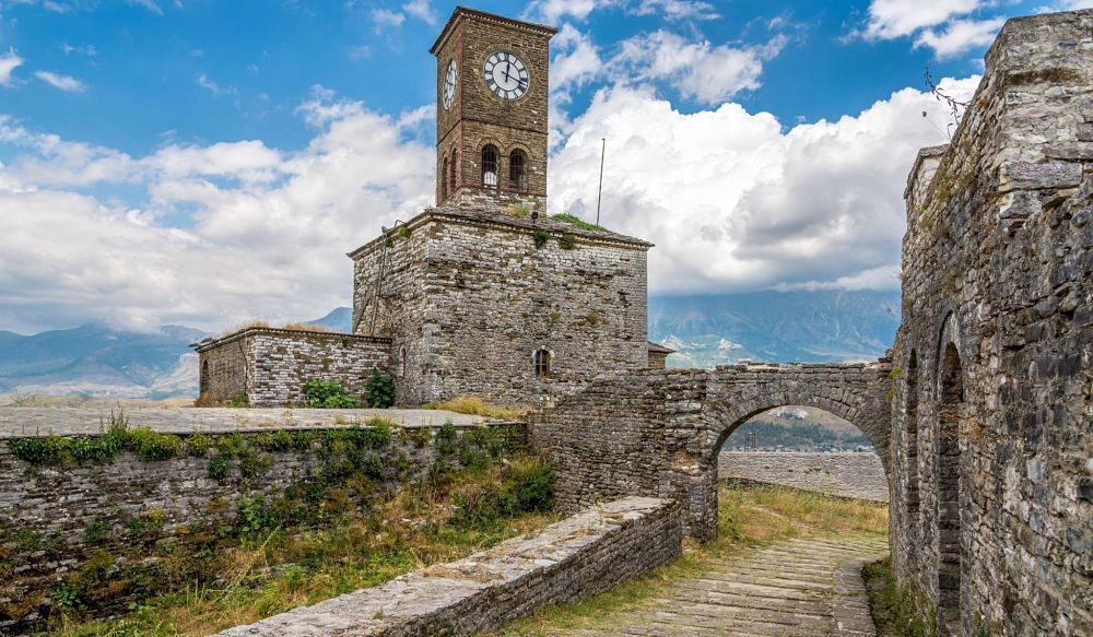 Une vieille tour de l'horloge en pierre sur une forteresse avec un pont en arc et des montagnes en arrière-plan sous un ciel partiellement nuageux.