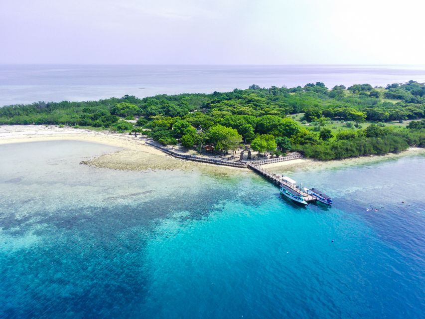 Eine Luftaufnahme einer kleinen tropischen Insel mit einem Holzsteg, an dem zwei Boote im türkisfarbenen Wasser festgemacht sind.
